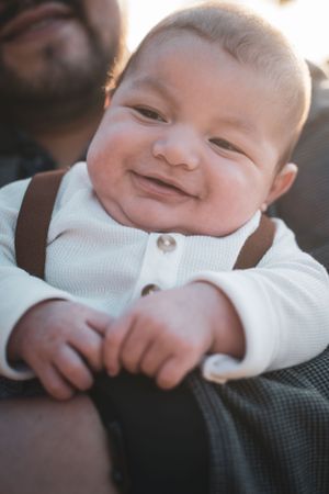 Adorable smiling newborn baby in white outfit with brown suspenders during family portrait.