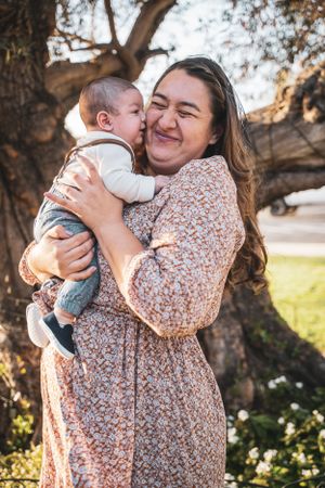Happy mother laughing while holding baby near a tree during golden hour family session.