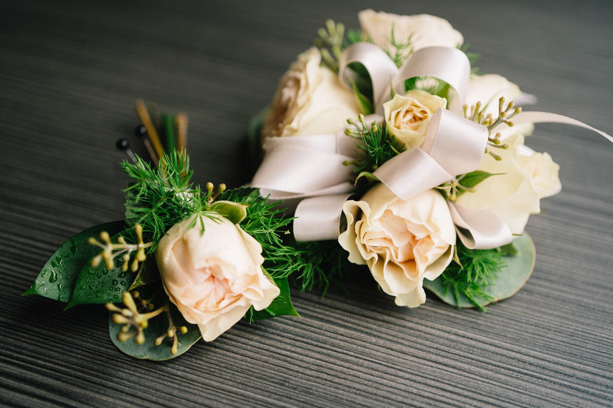 Soft peach and cream rose boutonnieres with greenery on dark surface.
