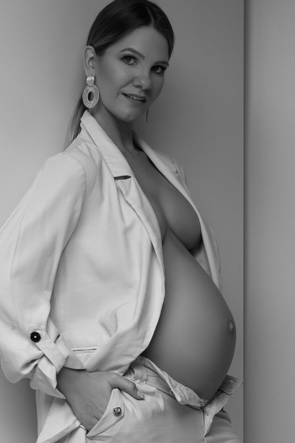 Artistic black and white maternity photo of an expectant mother in a white blazer against a light backdrop.