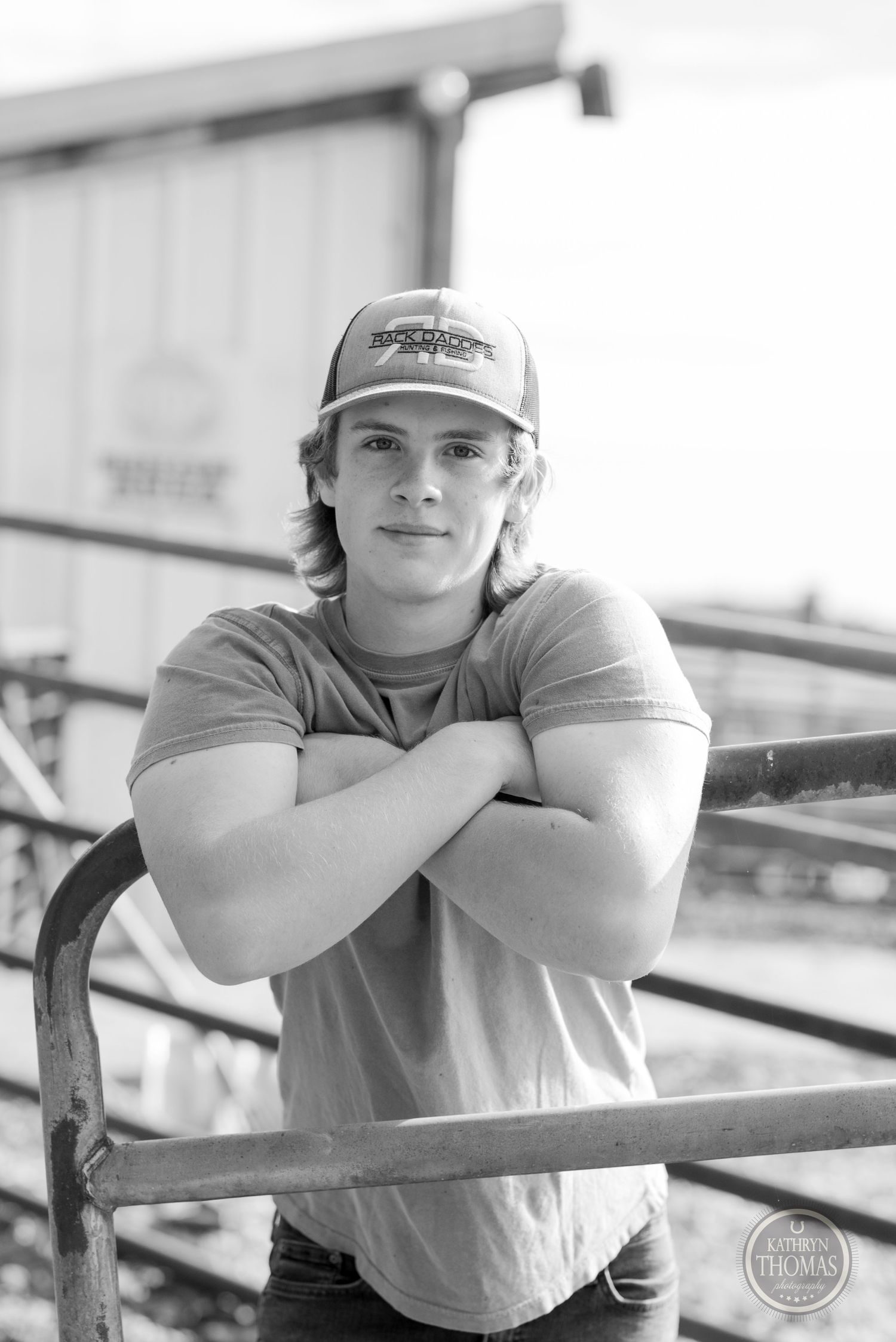 Farm worker in baseball cap leans on metal railing in black and white portrait.