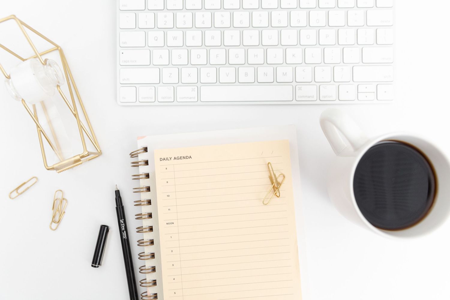 Minimalist desk setup with white keyboard, notebook, gold clips and coffee cup.