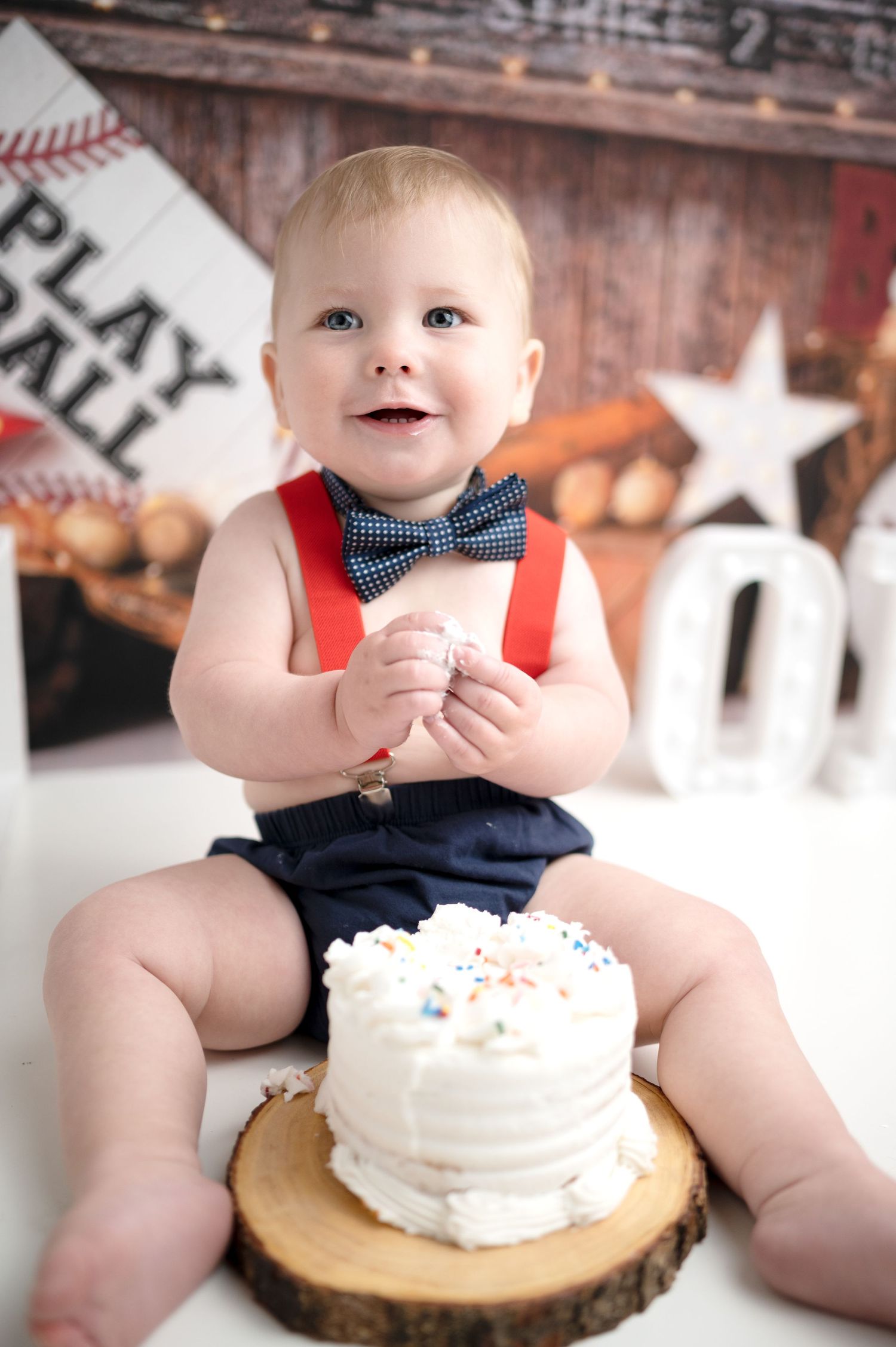 Baby in navy outfit with bow tie sits near a cake on a wooden slice with baseball themed decorations.