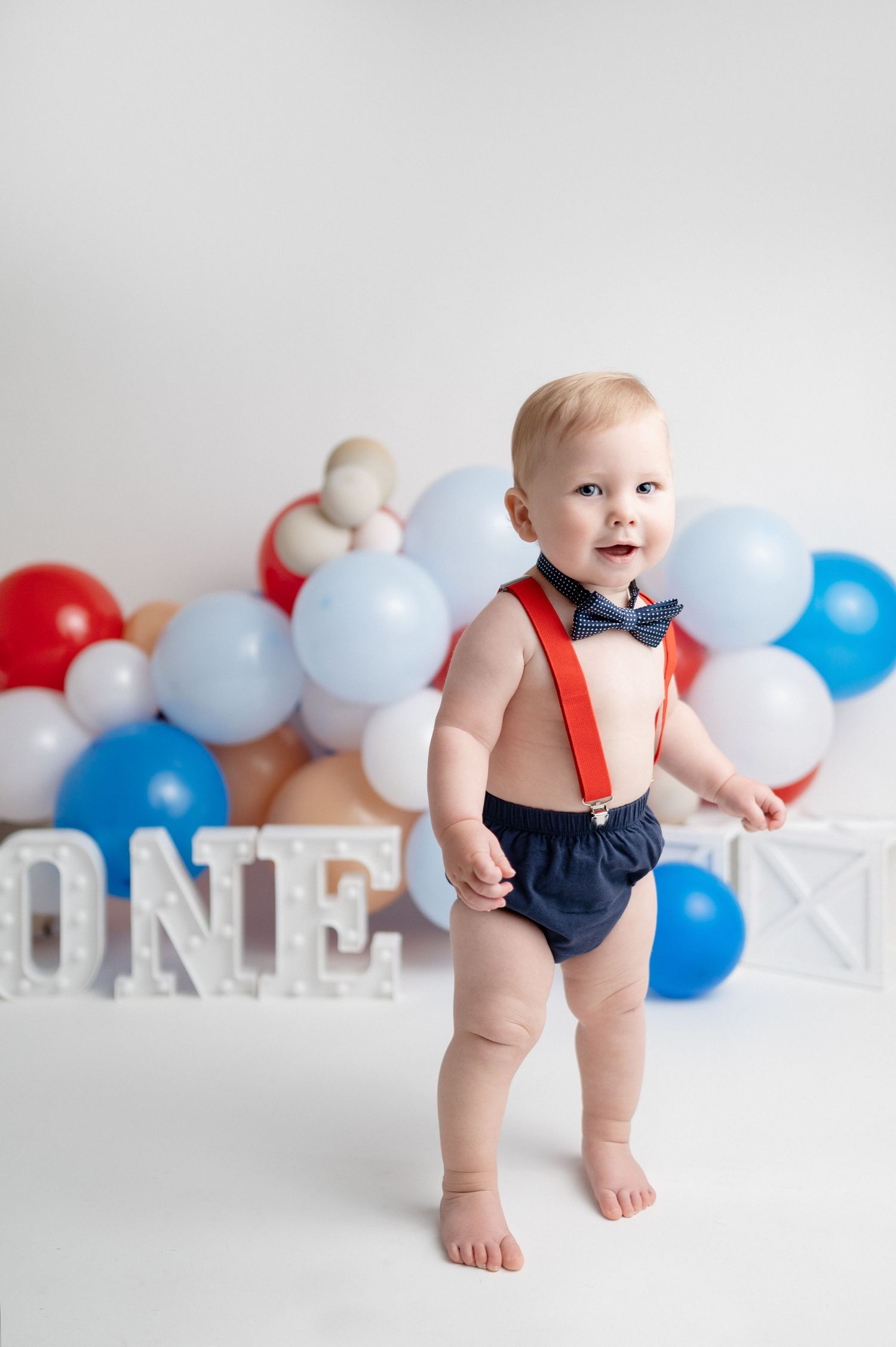 Smiling baby in suspenders and bow tie celebrates first birthday with balloons and decorations.