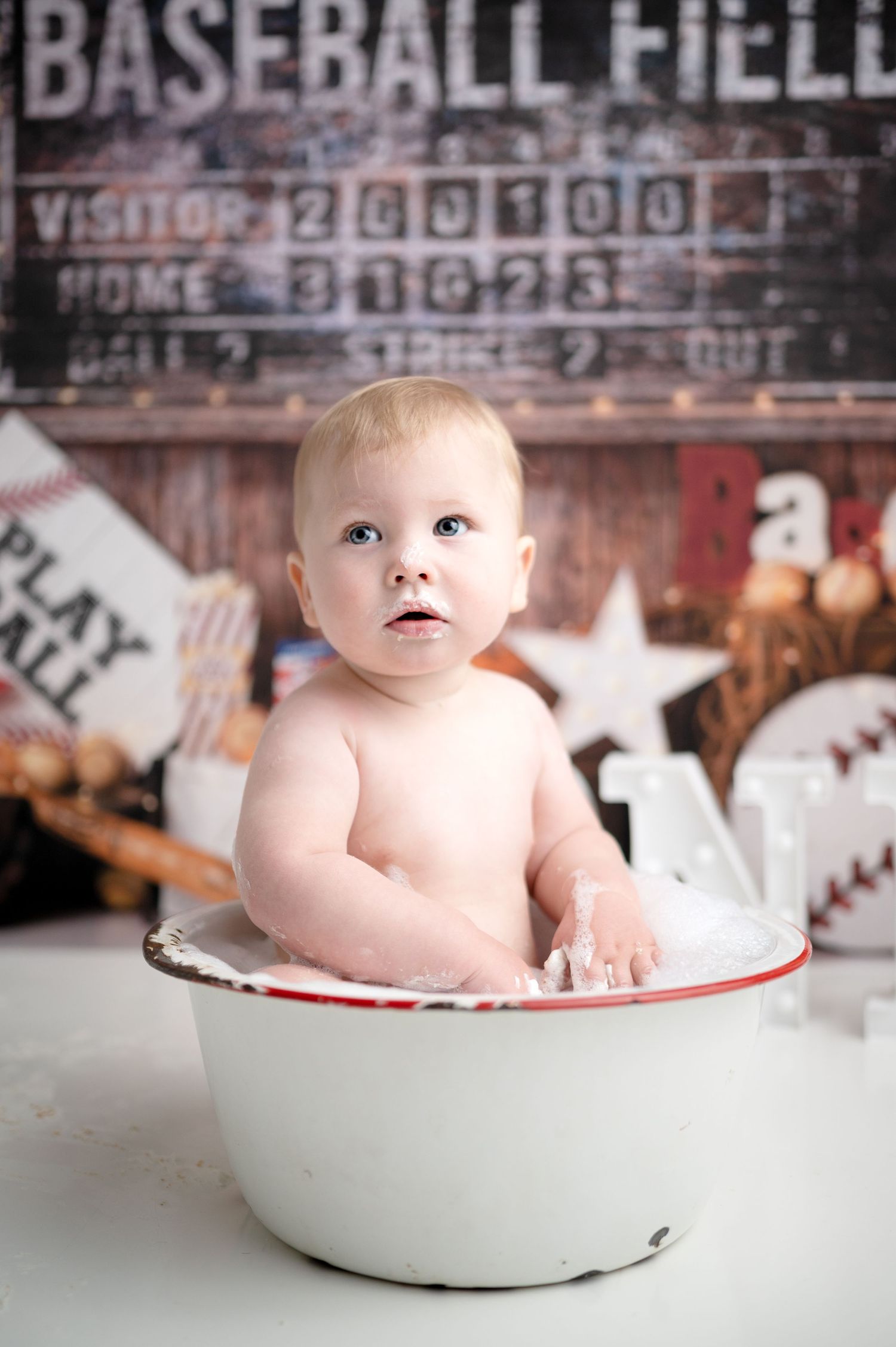 Baby sits in vintage white enamel basin against baseball themed backdrop for portrait session.