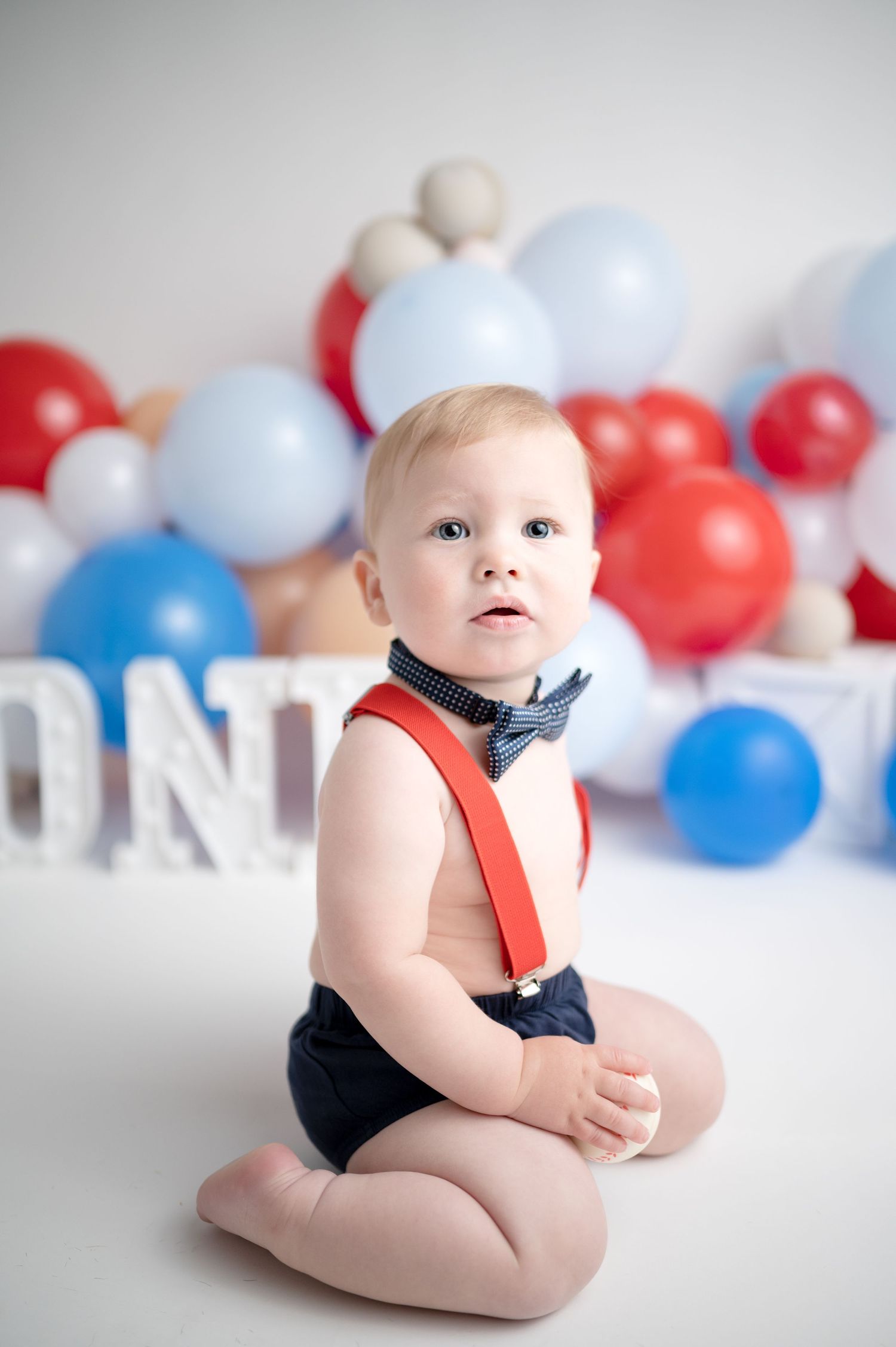 A baby in red suspenders and blue bow tie poses with red white and blue balloons in the background.