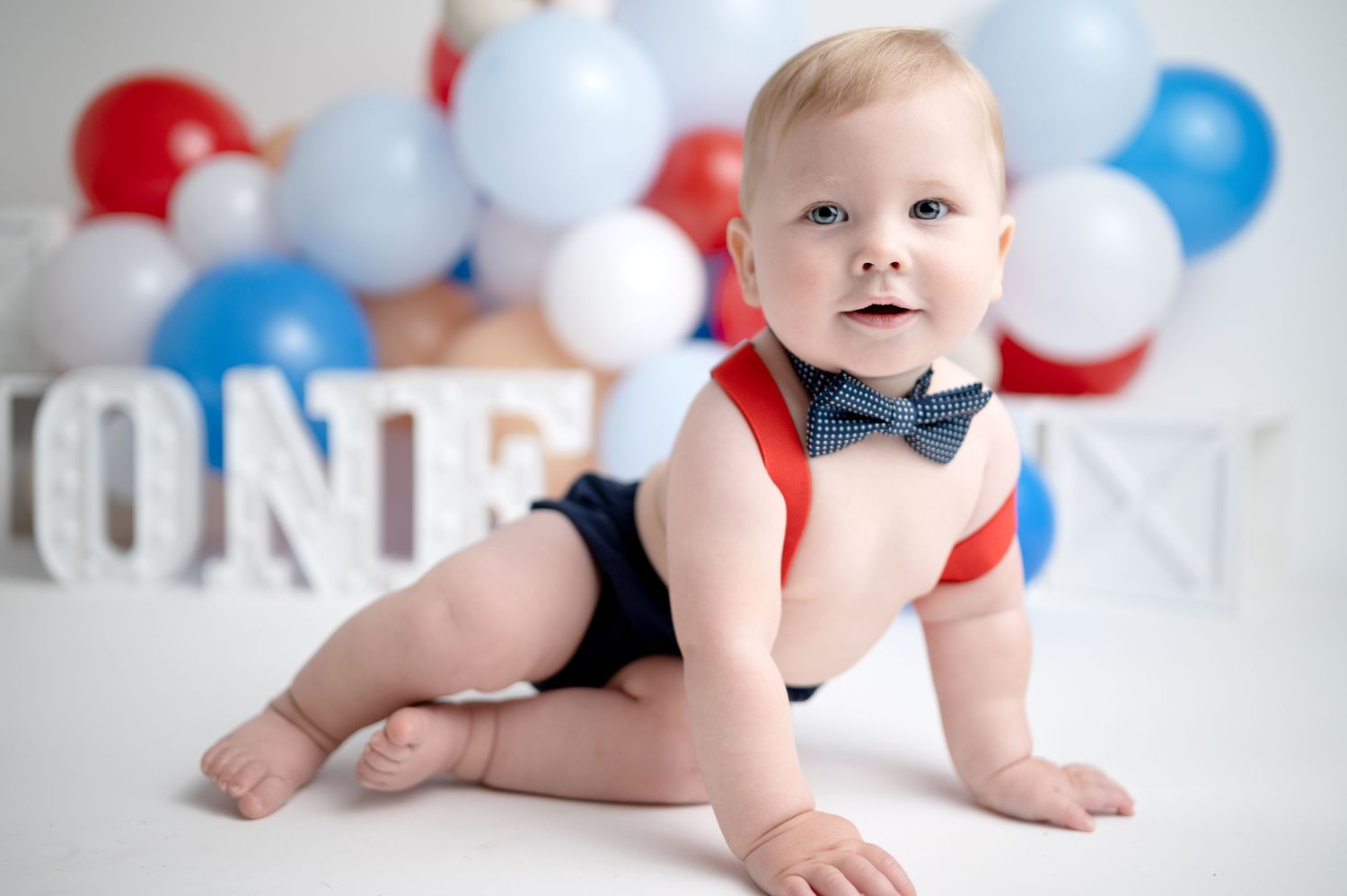 Baby in patriotic outfit sits with festive red, white and blue balloon decorations.