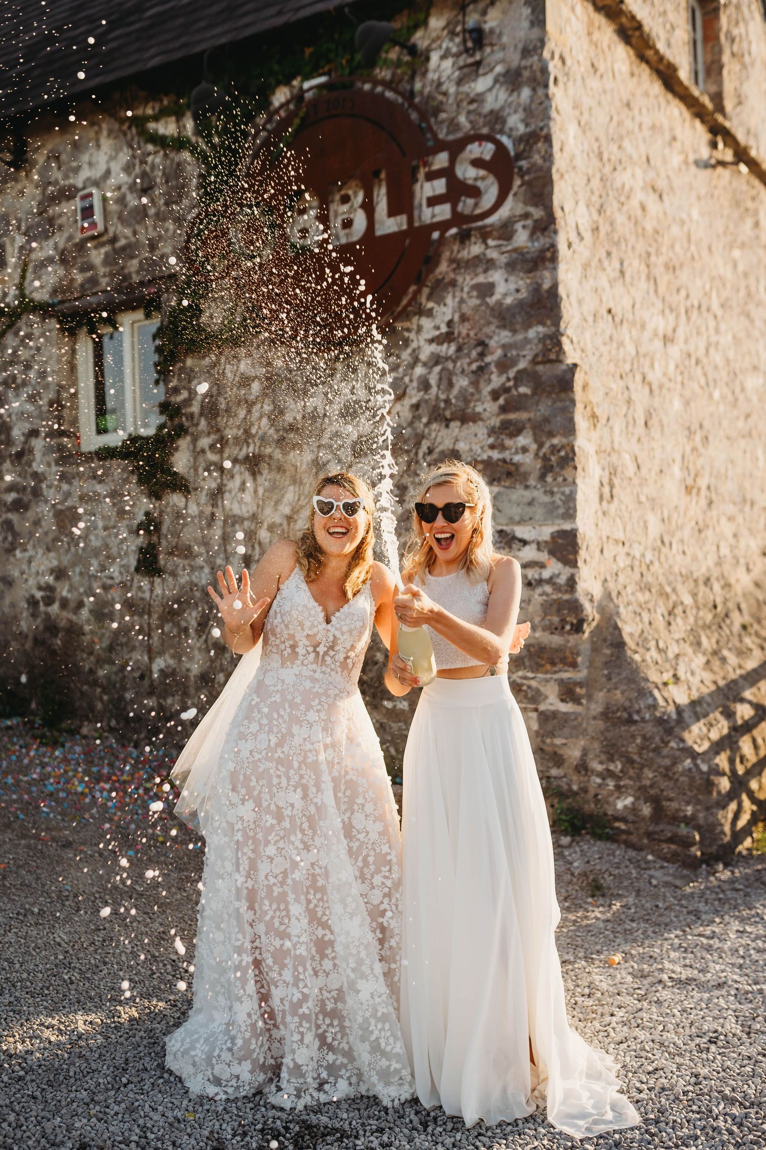 Two brides in white gowns and sunglasses celebrate with champagne spray outside Cobbles Wedding Venue Ogmore, South Wales