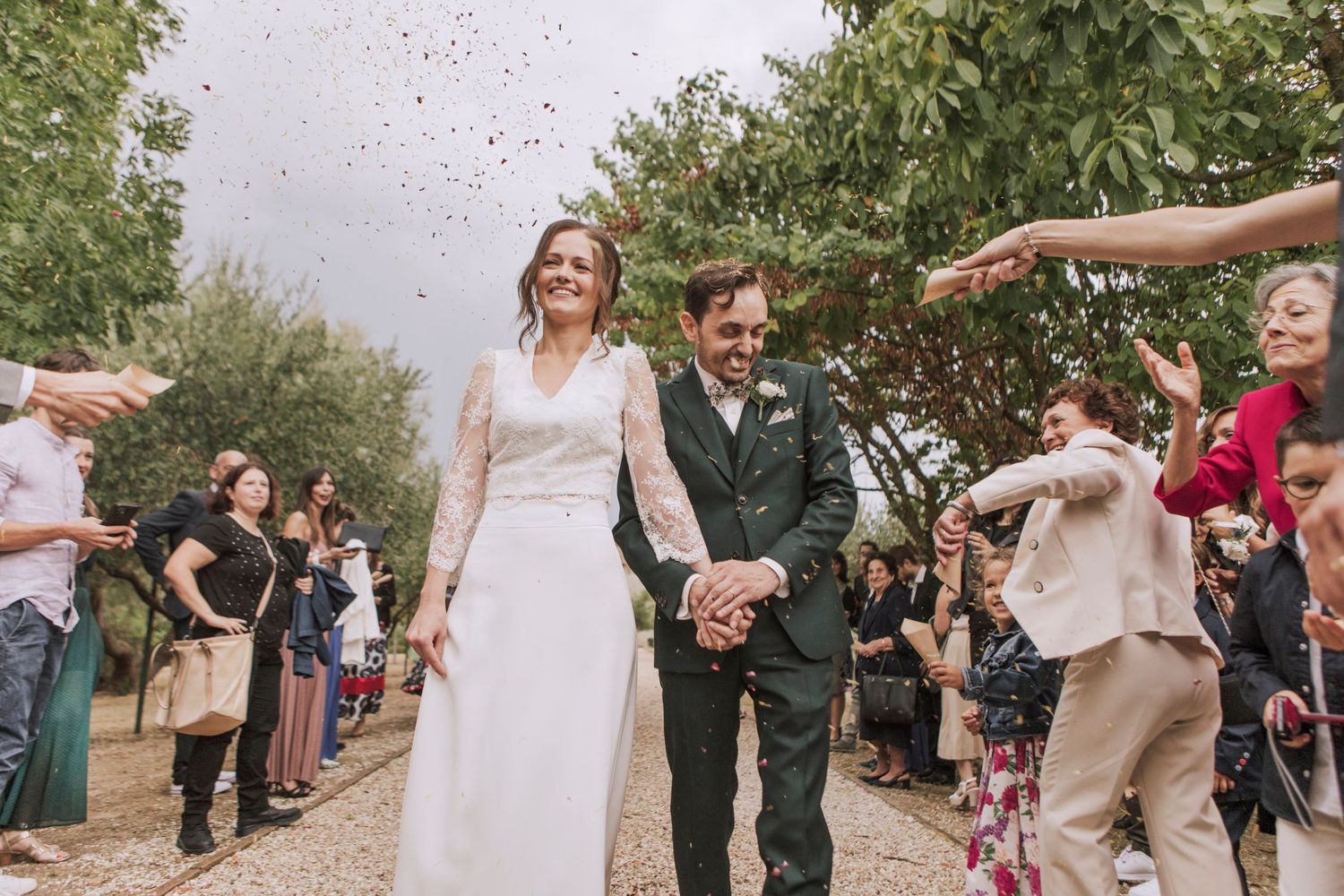 Newlyweds walking through crowd of guests throwing confetti on tree-lined path.