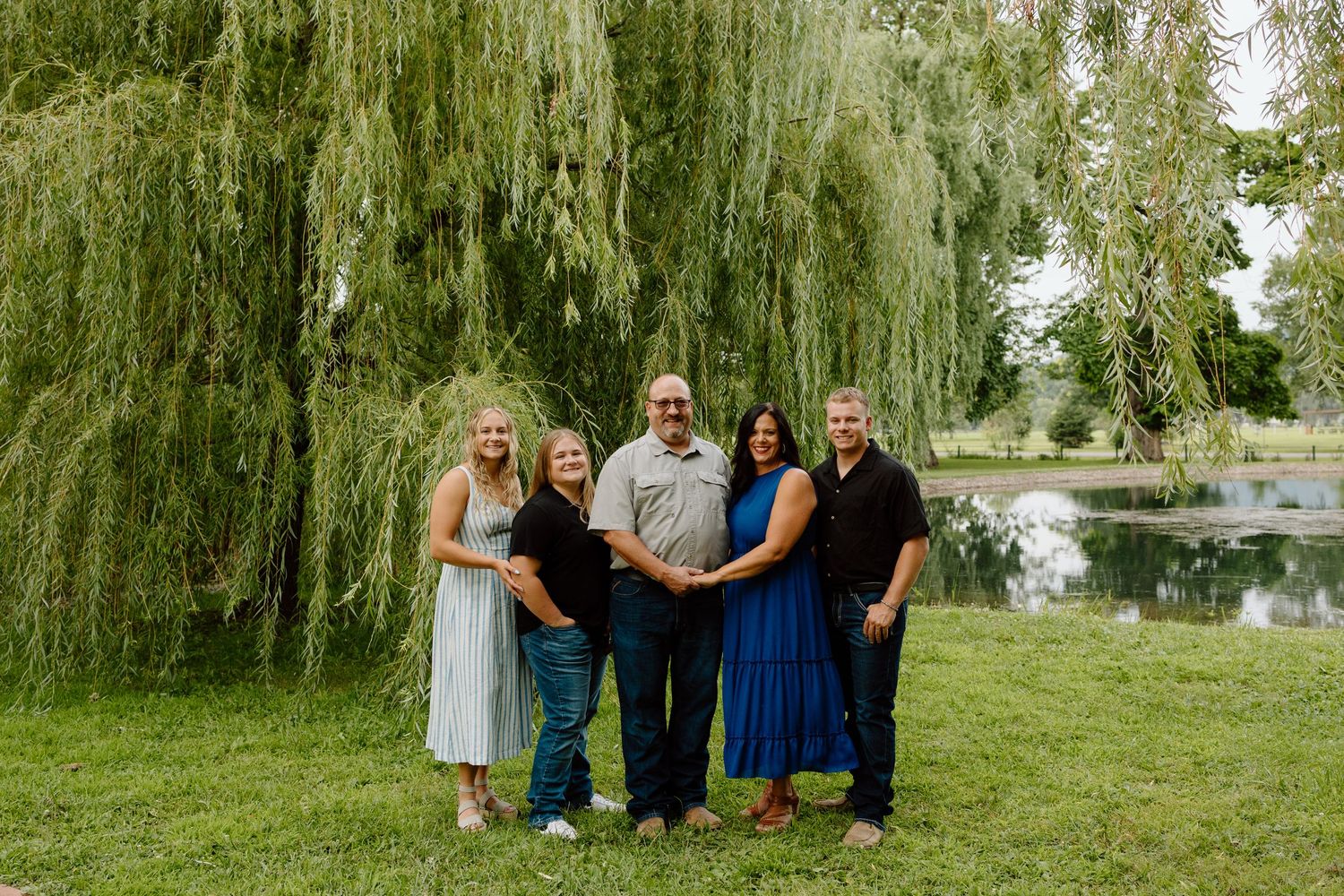Adult family of five smiles together on green grass beneath a weeping willow by a pond.