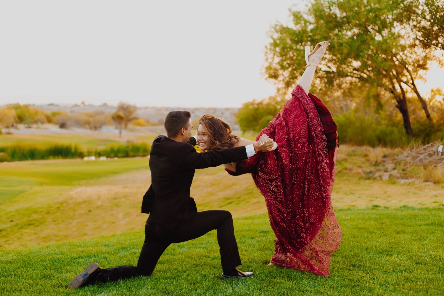 Couple in formal attire dancing dramatically on grass during golden hour.