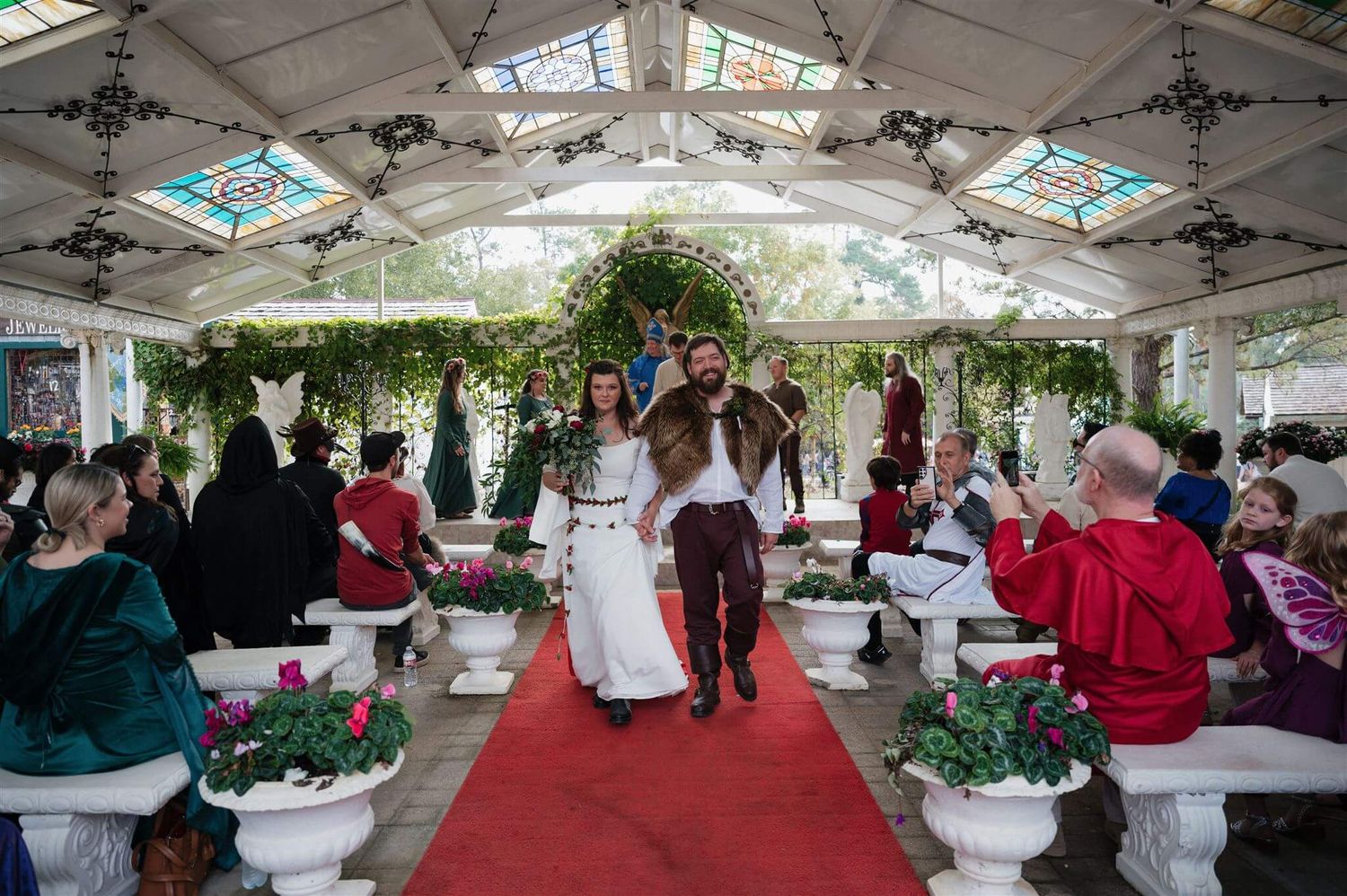 A medieval wedding reception in a decorated pavilion with guests seated along a red carpet aisle.