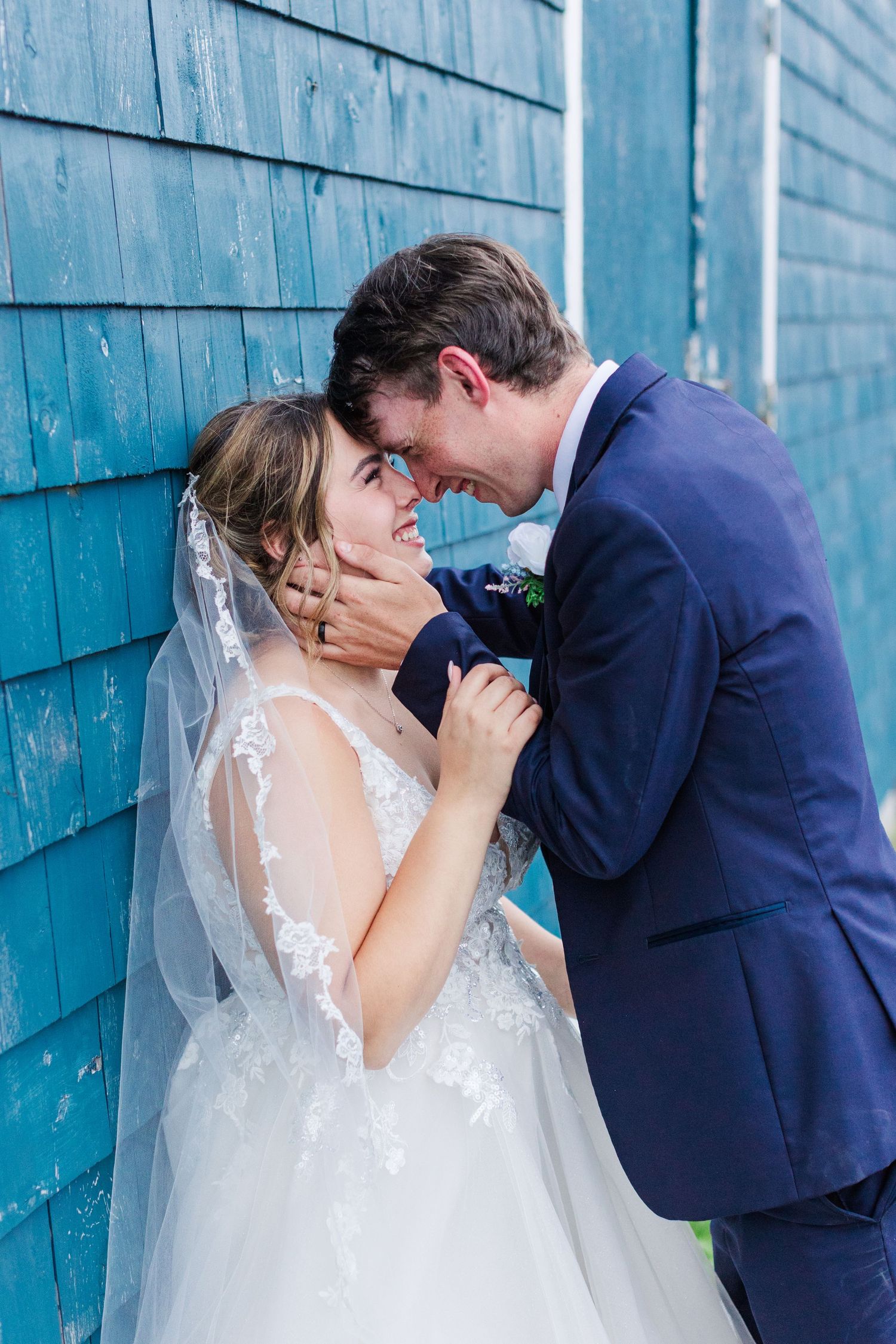 Newlyweds share an intimate moment against a blue wooden wall backdrop while dressed in wedding attire.