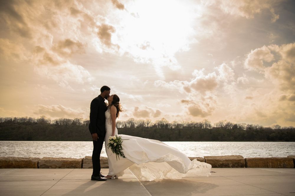 Silhouetted wedding couple embracing at sunset by a waterfront with dramatic sky.