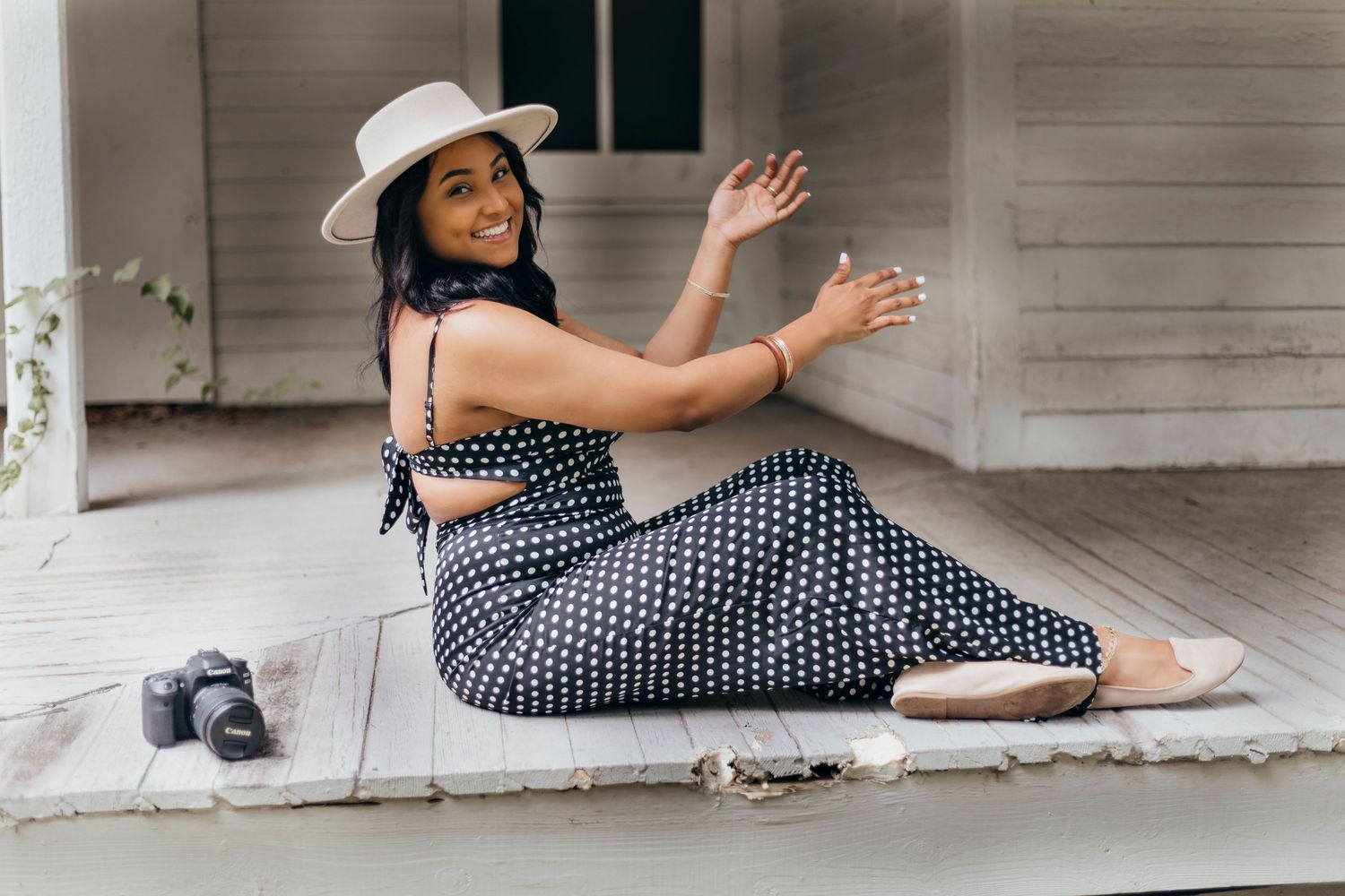 Photographer in polka dot dress and white hat poses creatively on white wooden porch.