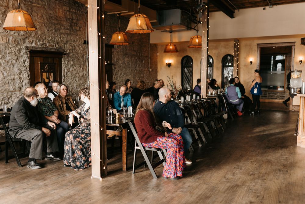 Wedding guests seated in rows inside a rustic stone venue with warm pendant lighting.