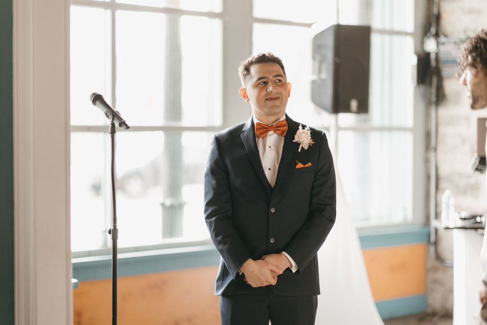 Groom in navy suit and orange bow tie waiting emotionally at altar during wedding ceremony.