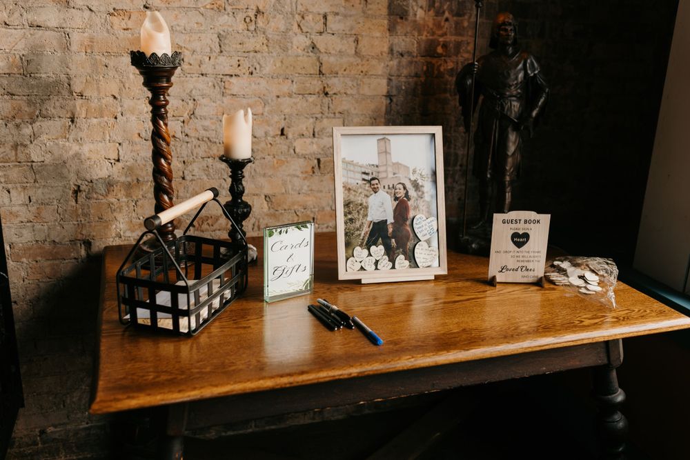 Rustic wedding guest book table with candles, framed photo, and pen on wooden surface against brick wall.