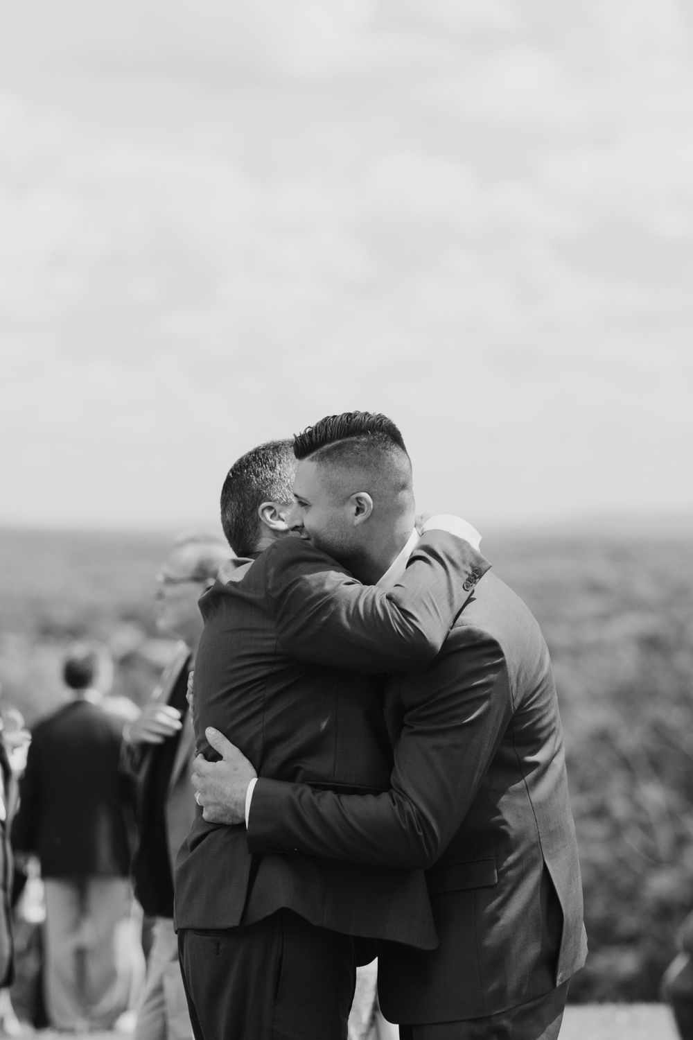 Two men in suits share an emotional embrace at an outdoor wedding.