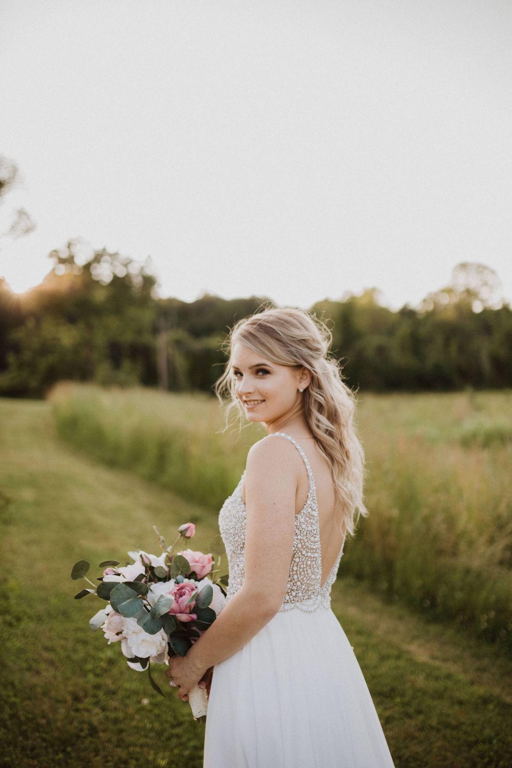 Bride in a lace wedding gown holds bouquet while posing in a sunset field.