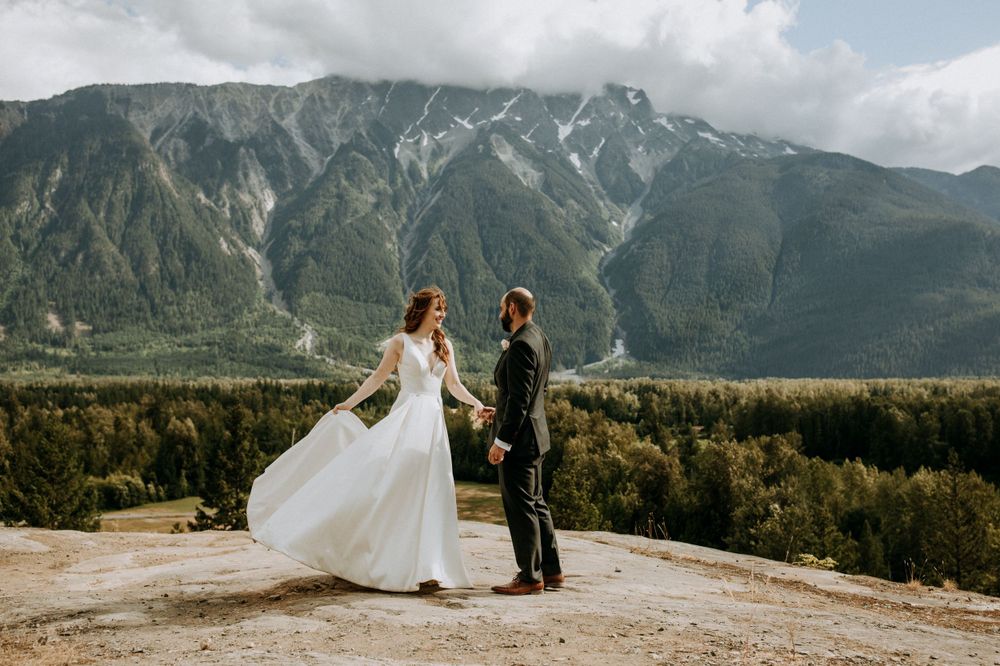 A couple in wedding attire stands on a mountain ridge overlooking a dramatic mountain range and forest landscape.