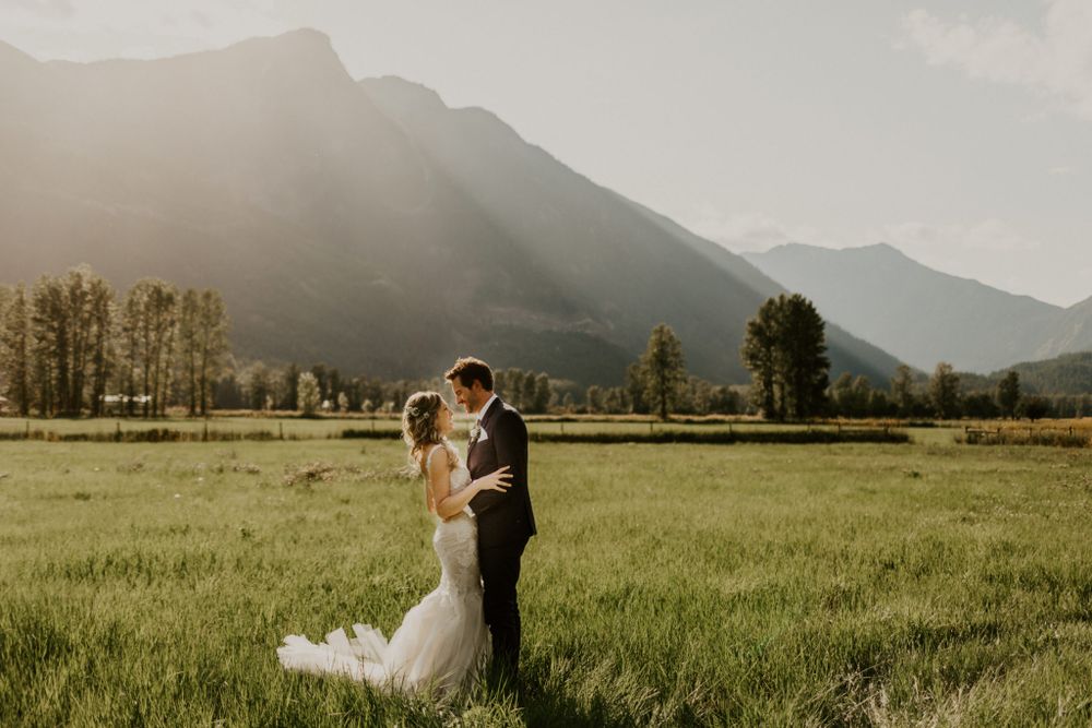 A couple embraces in a meadow with majestic mountain peaks and sun rays filtering through clouds in the background.