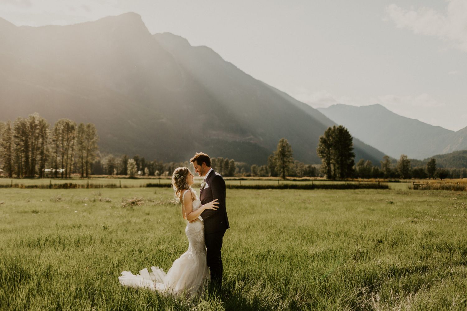 A couple embraces in a meadow with majestic mountain peaks and sun rays filtering through clouds in the background.