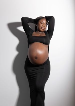 Artistic maternity portrait of an expectant mother in black attire against a white backdrop with dramatic shadow.