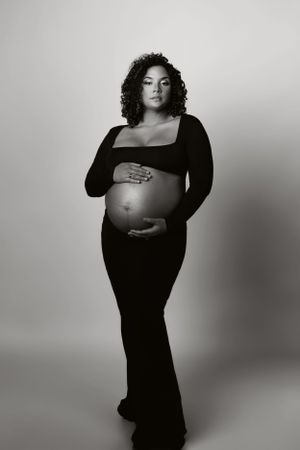 Black and white maternity portrait featuring woman in long dress against neutral background.