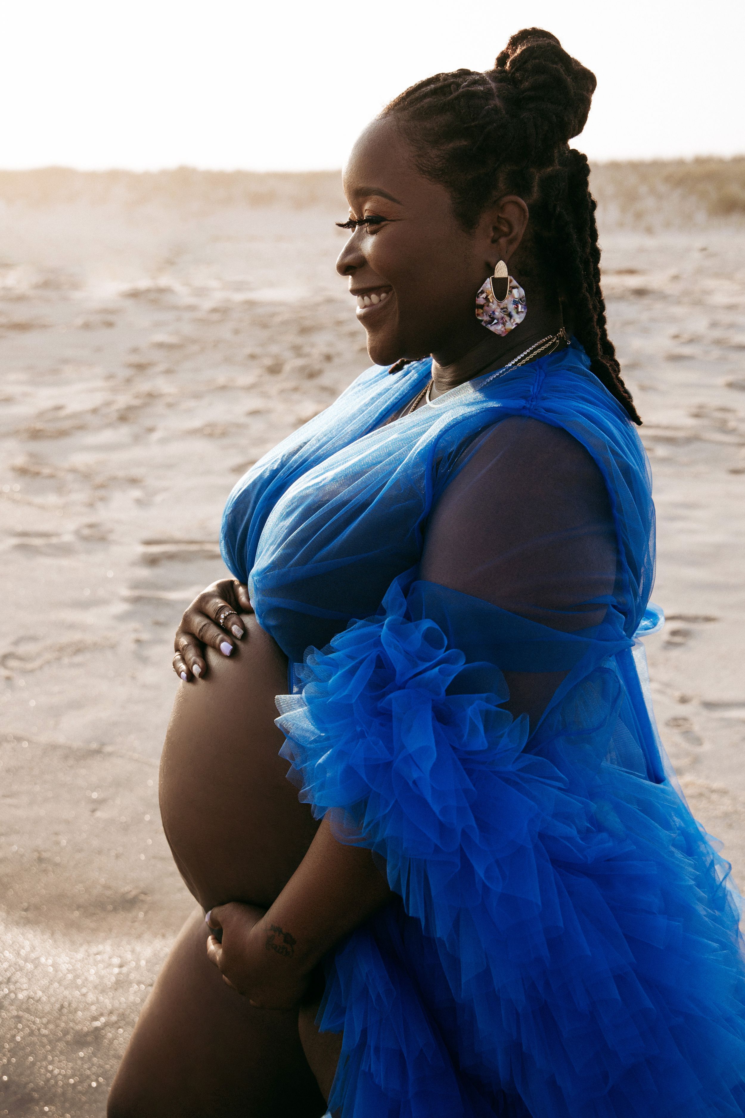 Expectant mother wearing royal blue tulle dress smiling on beach at sunset.