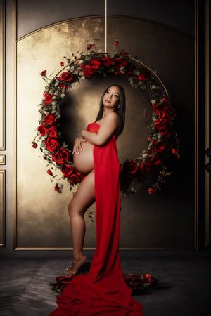 Artistic maternity photo with red roses forming a circular arch and flowing red fabric.