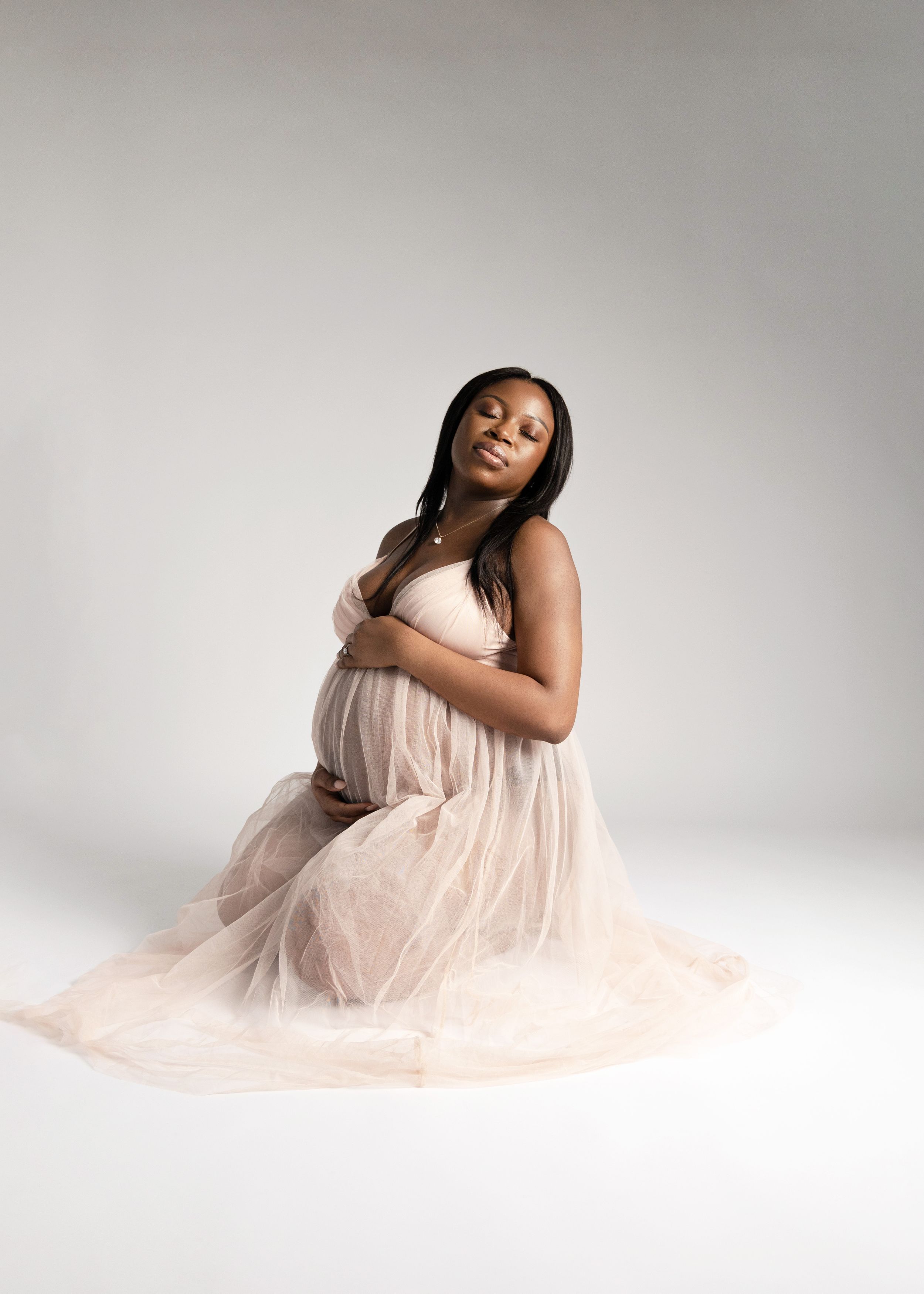 Elegant maternity portrait with woman in flowing blush tulle dress against white backdrop.
