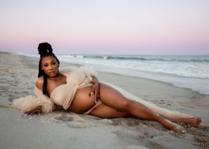 Expectant mother in beige dress reclining on beach at sunset during maternity photoshoot.