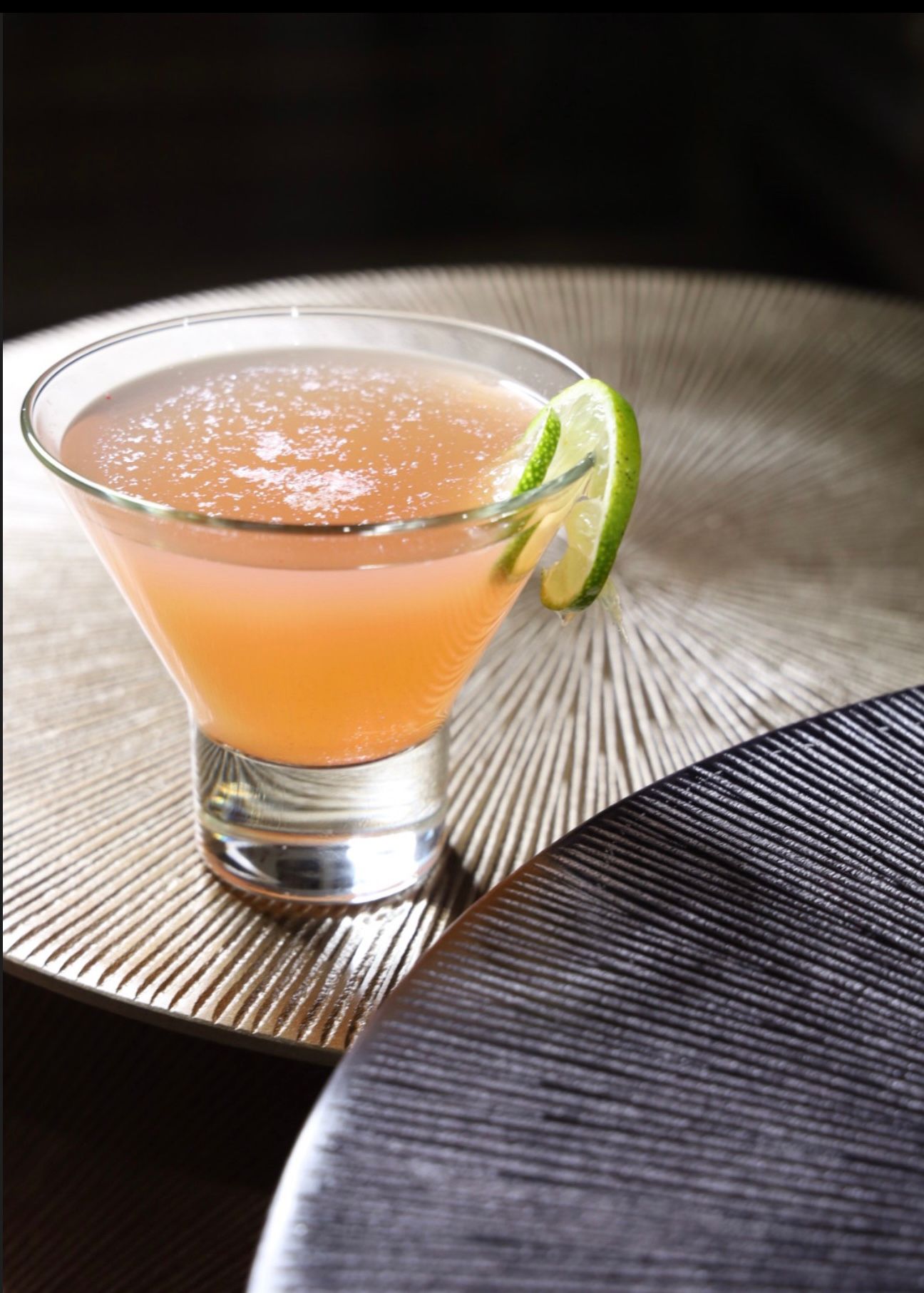 Pink cocktail with lime garnish served in martini glass on textured surface.