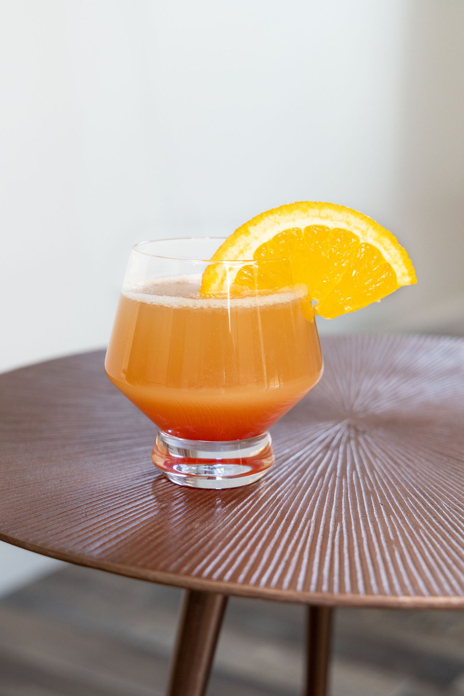 Orange-colored cocktail with citrus garnish served in coupe glass on textured table.