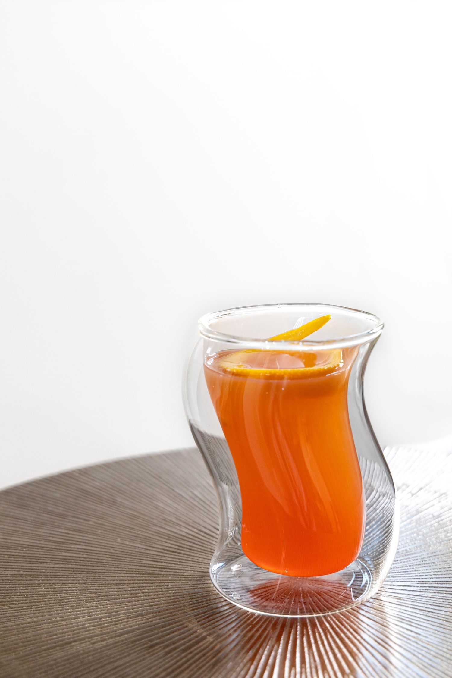 An orange cocktail served in a curved double-walled glass with citrus garnish.