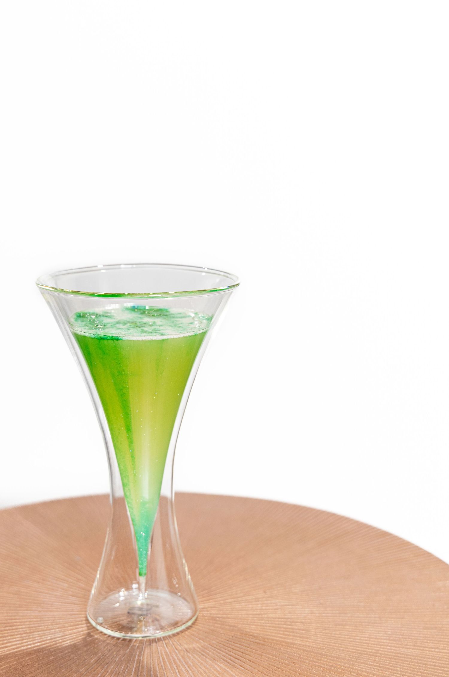 Bright green cocktail served in hourglass-shaped martini glass on wooden surface.