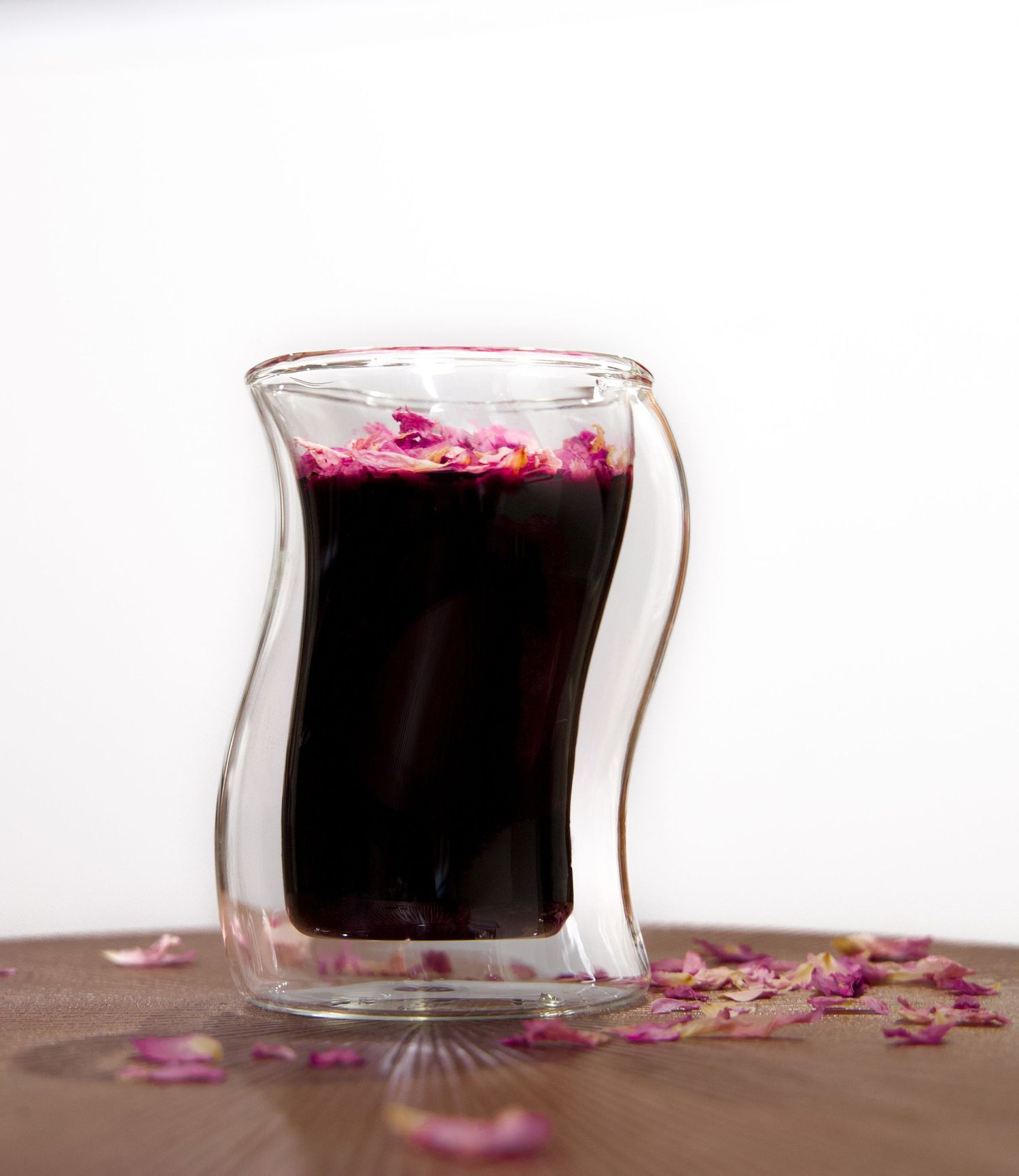 A dark purple beverage in a double-walled glass cup garnished with pink flower petals.