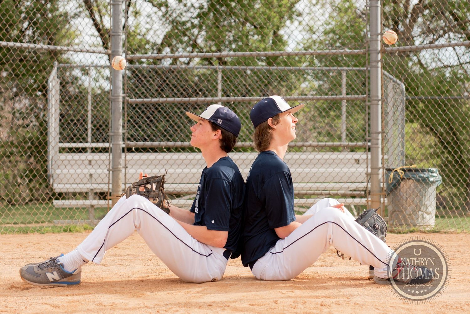 Baseball teammates in uniform sitting back to back on dirt field during practice session.