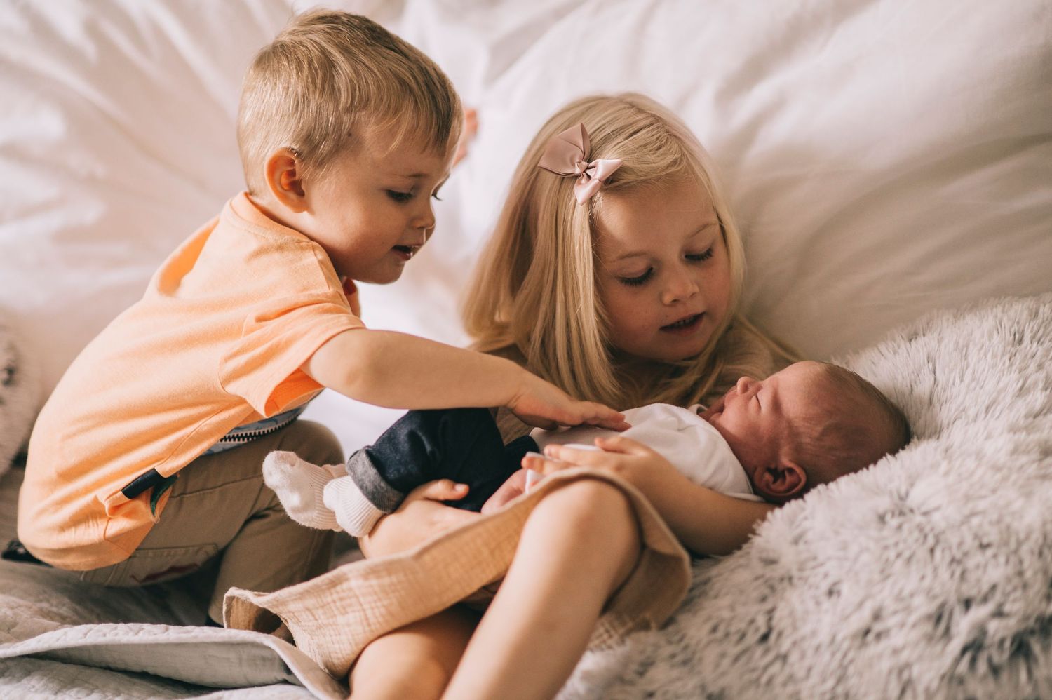 Two young siblings tenderly hold their newborn baby brother on a cozy white bed.