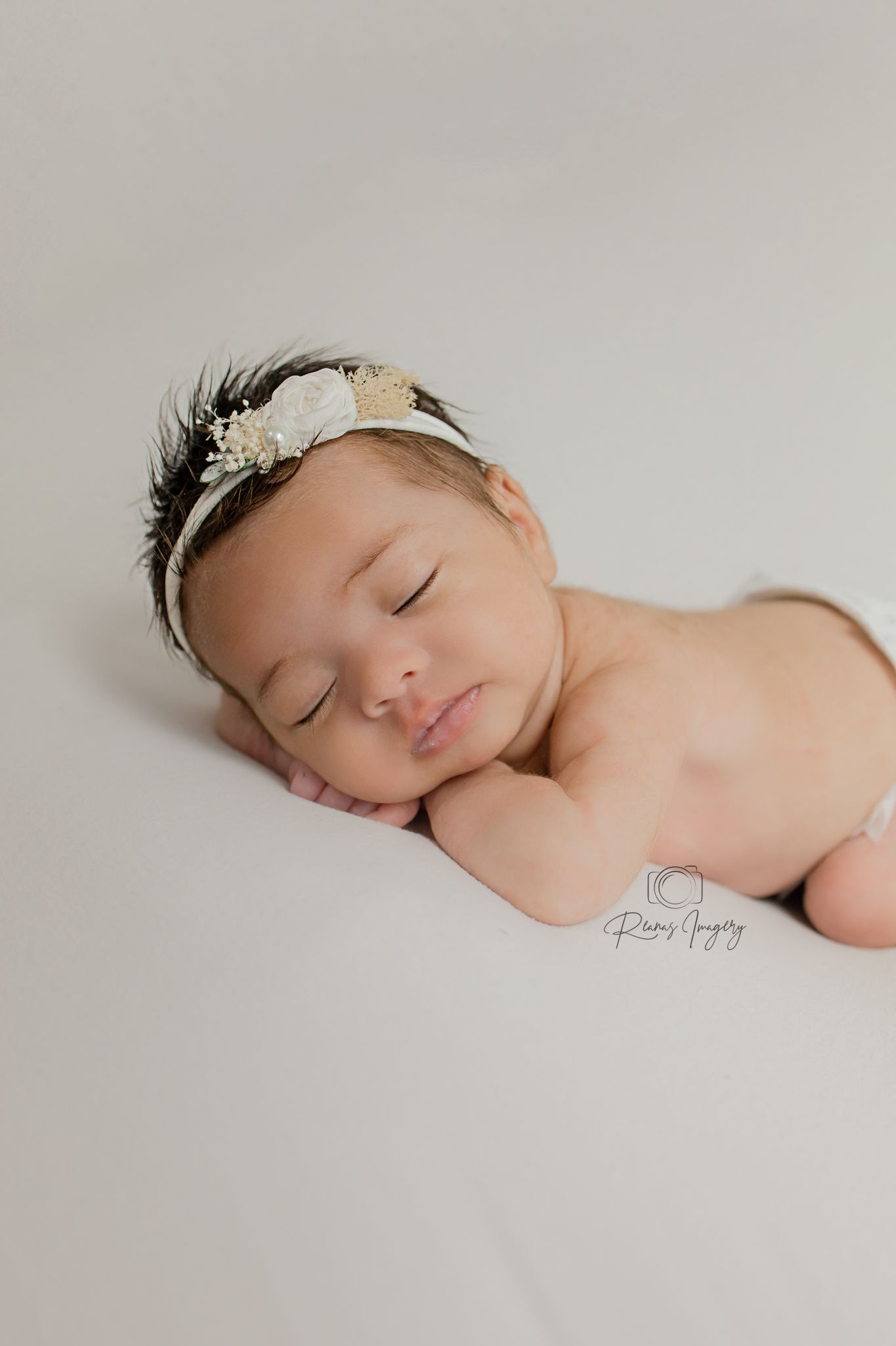 Peaceful sleeping newborn baby wearing a decorative feather and floral headband on cream backdrop.