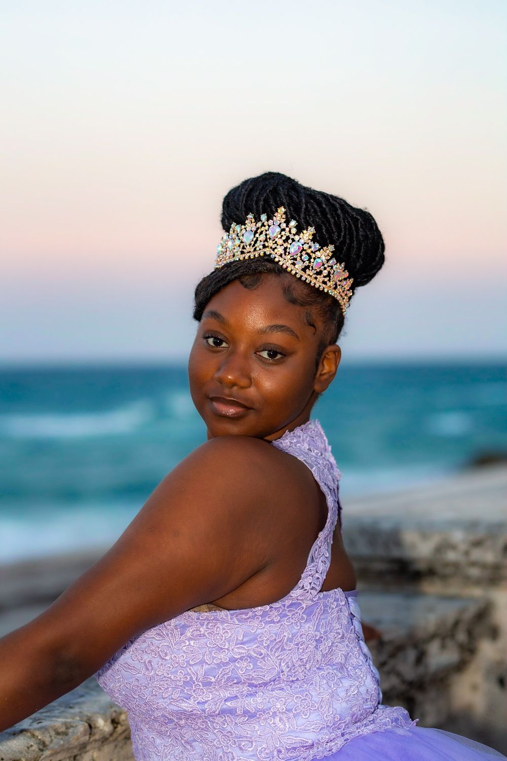 Portrait at beach sunset wearing purple gown and crystal crown against ocean backdrop.