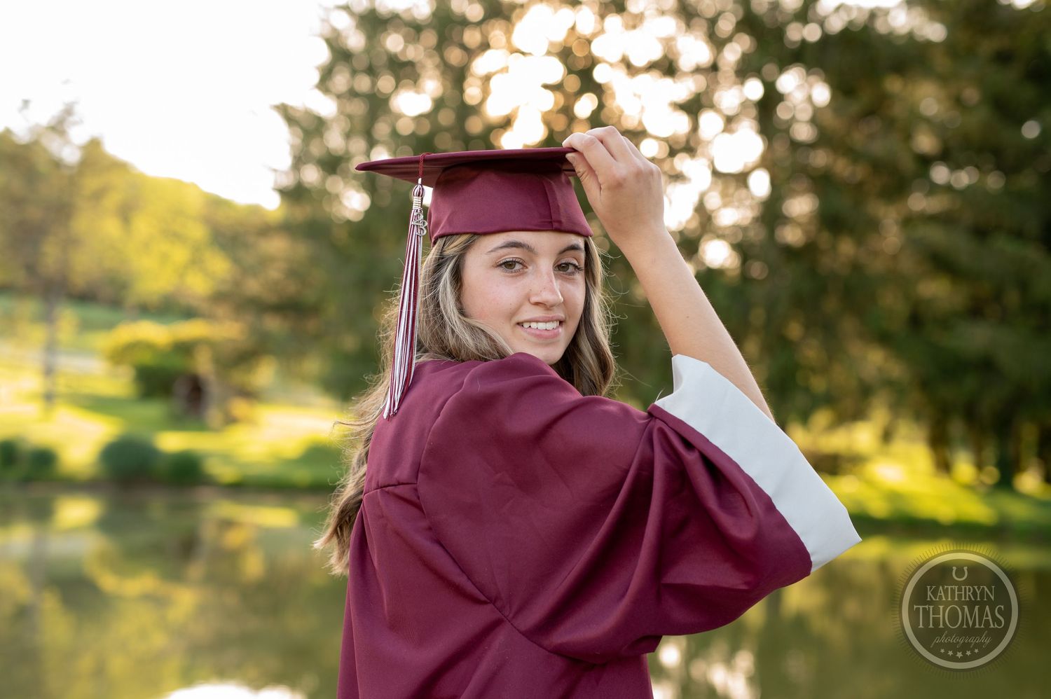 Graduate in burgundy cap and gown adjusts tassel outdoors near pond.