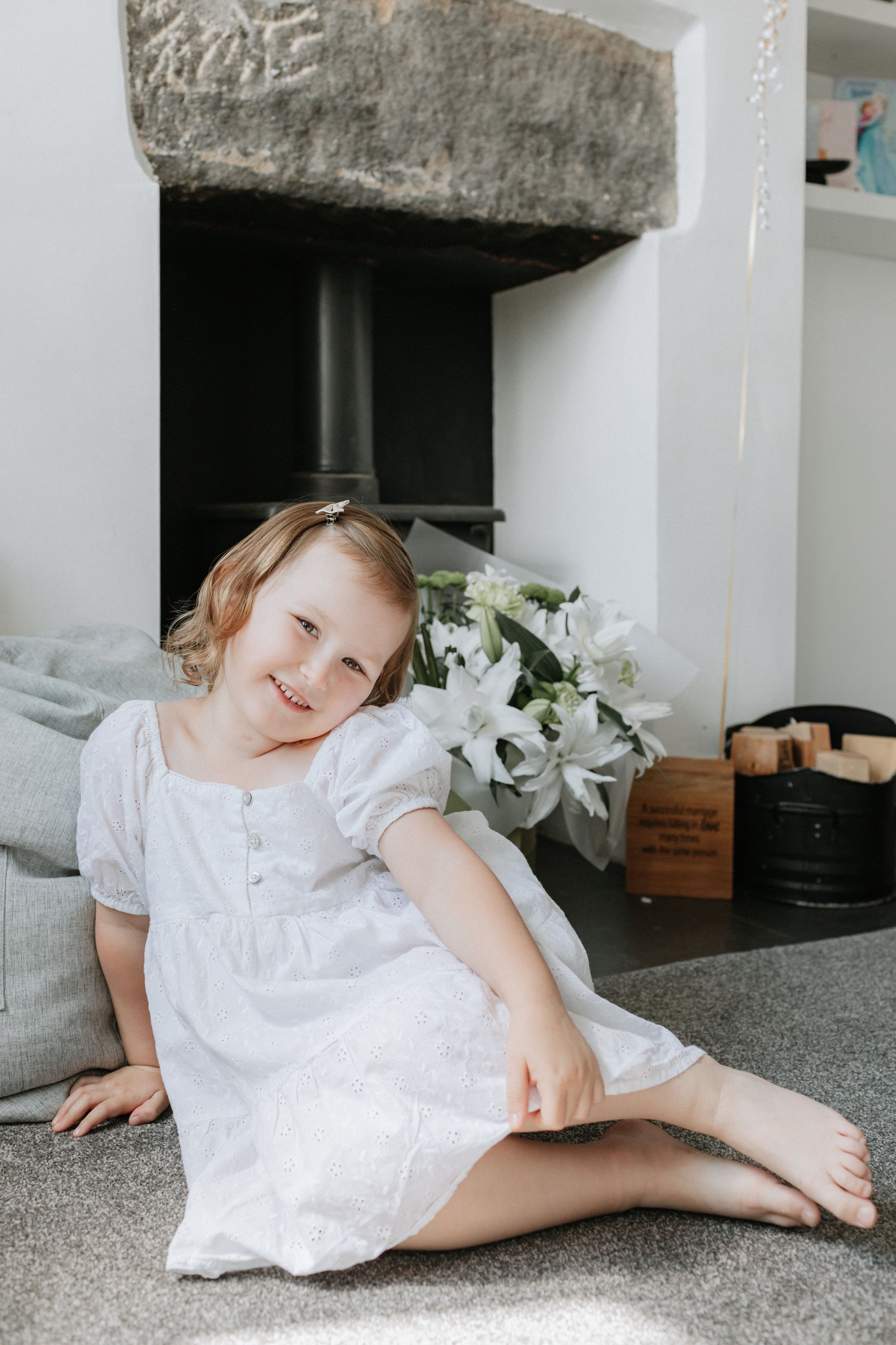 Young child in white dress sitting on carpet near a fireplace with white flowers.