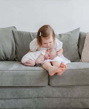 A young child in a white dress lounges playfully across a gray couch.