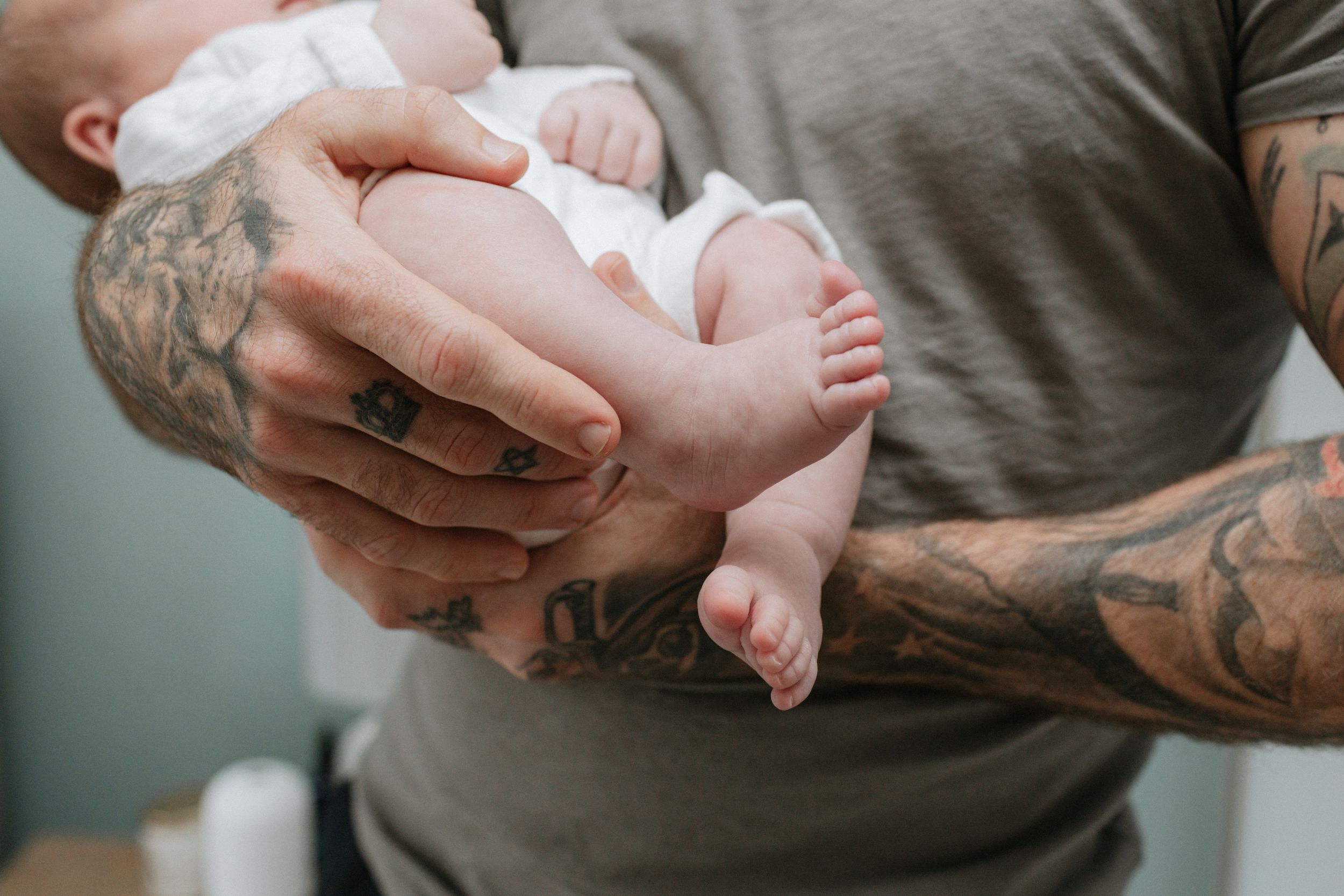 Tattooed hands gently holding tiny baby feet against gray shirt.
