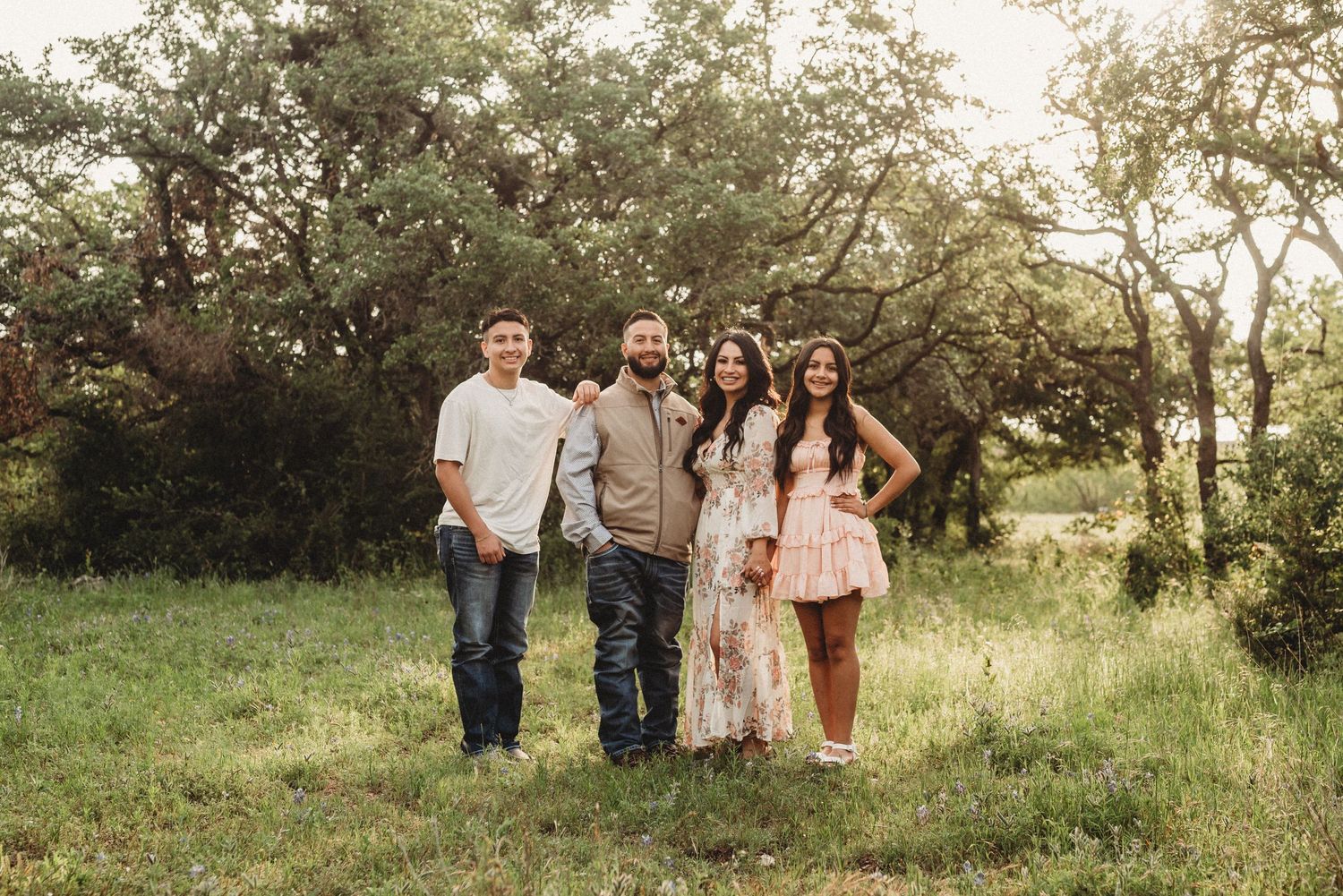 Group portrait standing together on grassy hill during golden hour sunset with trees in background.