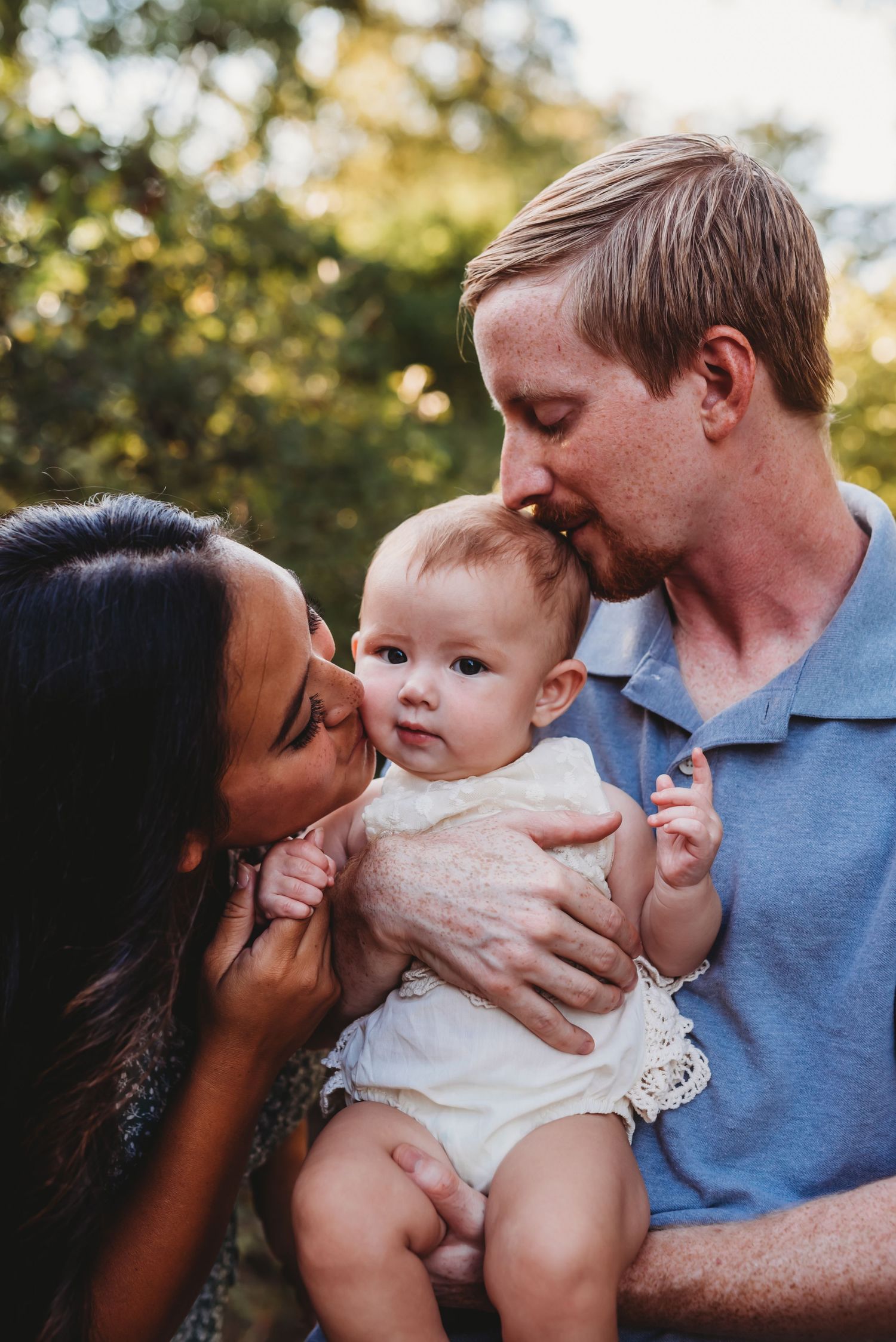 A tender family moment as parents share affection with their baby outdoors in warm sunlight.