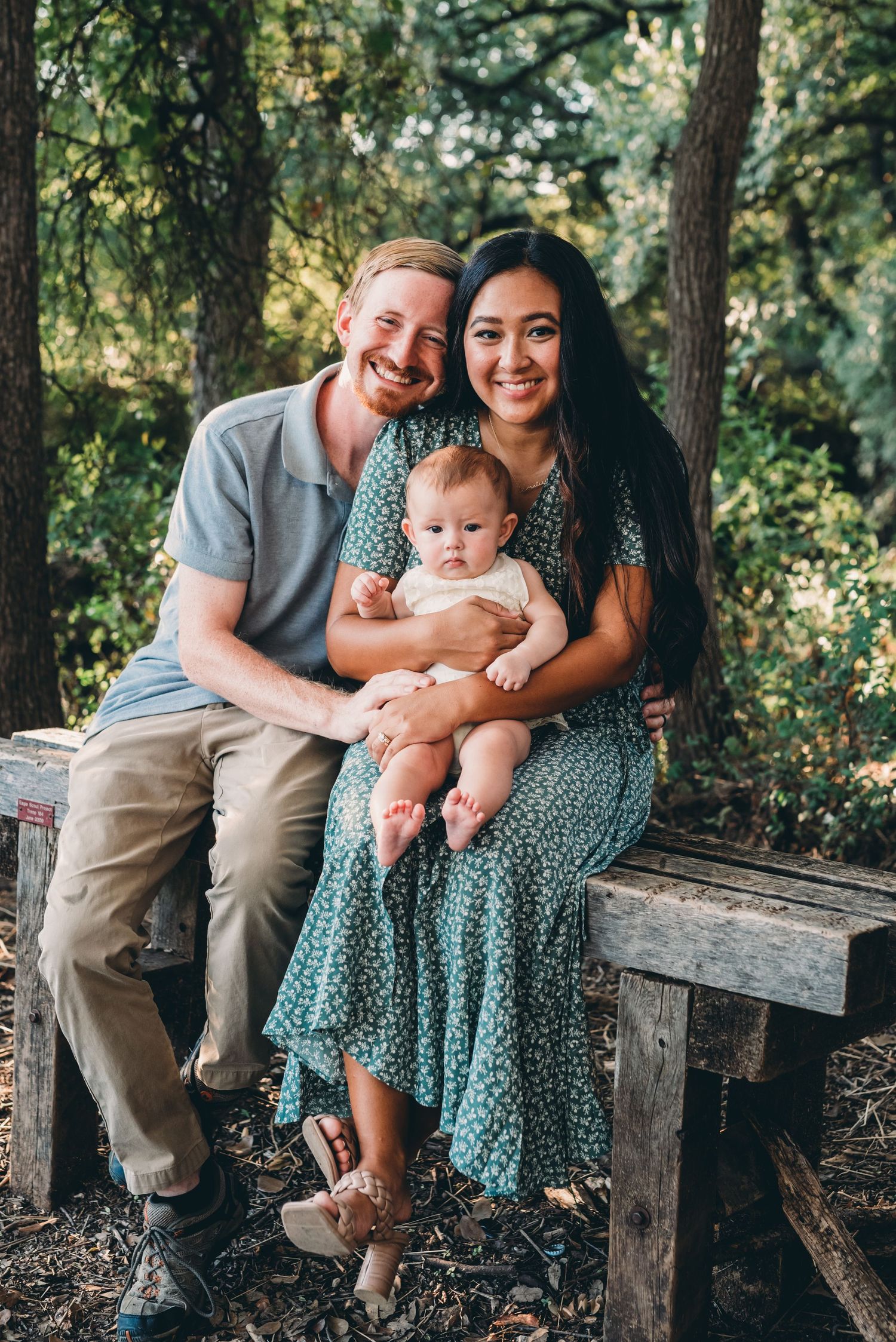 Family sitting together on wooden bench in wooded setting.