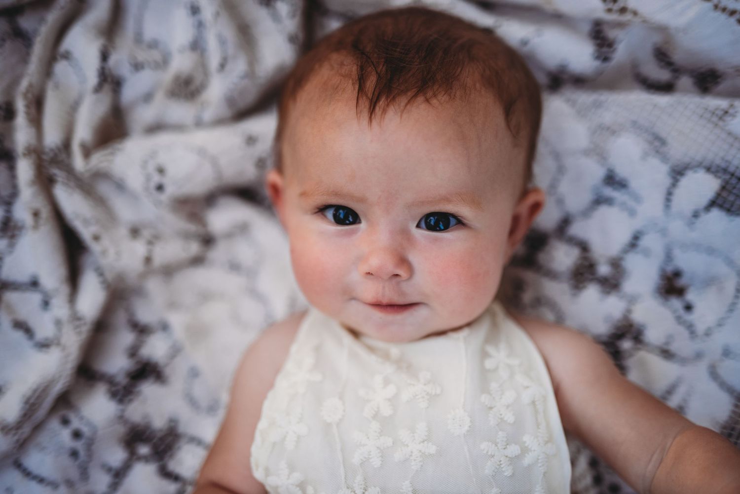 Baby wearing white lace dress smiles while lying on gray patterned blanket.